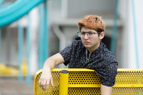 A portrait of a 20-year-old man wearing a short sleeve button up shirt and glasses leaning over the back of a yellow park bench.