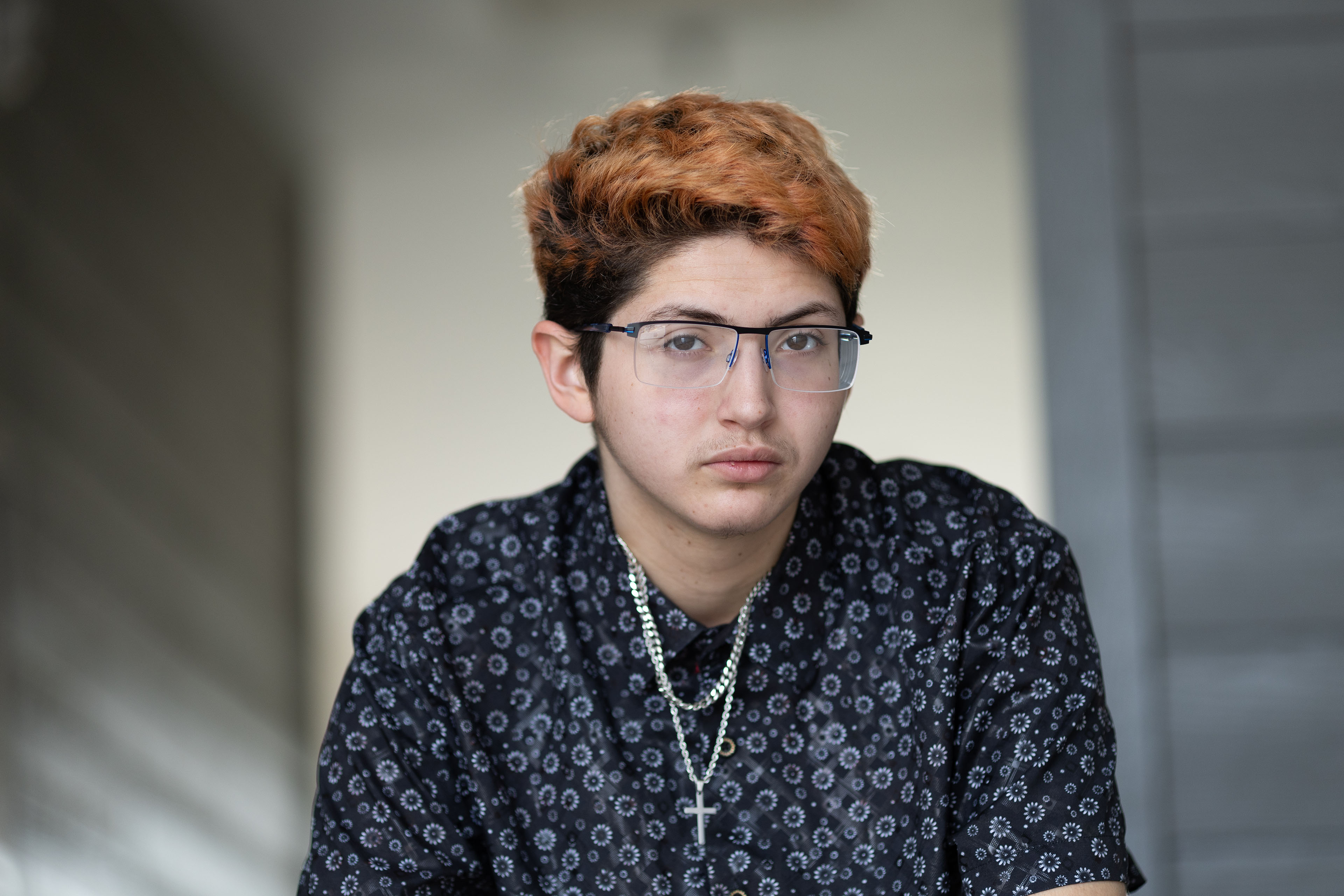 A portrait of a 20-year-old man. He wears glasses and a button-down short sleeve shirt. A silver cross necklace hangs from his neck.
