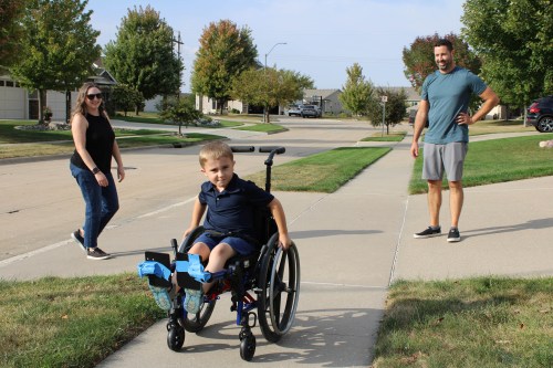 A young child in a wheelchair plays in a sidewalk with his mother to his left and his father to his right.