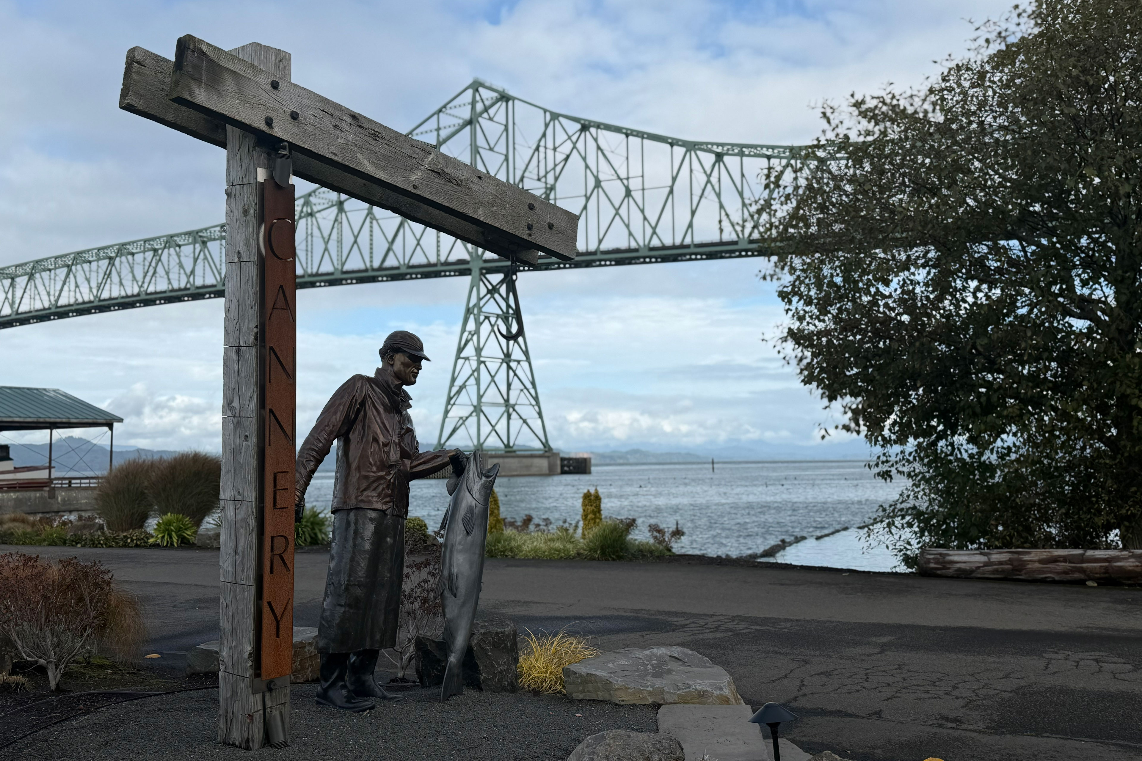 A statue of a woman holding a large fish is placed next to a sign reading "Cannery" at Astoria's waterfront. A bridge over the river is behind.