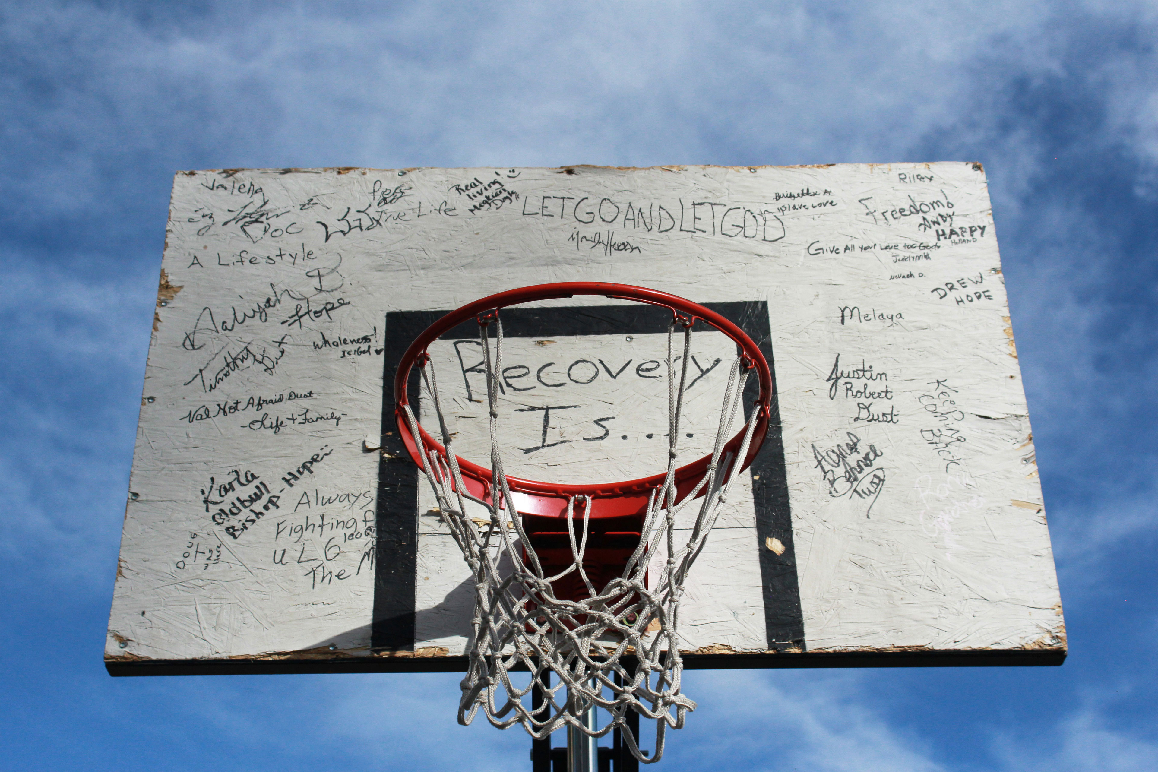 The weathered backboard of a basketball hoop is covered in handwritten messages. Directly behind the hoop reads "Recover is..." and examples of surrounding writings are "Freedom!," "Let go and let God," and "Hope."