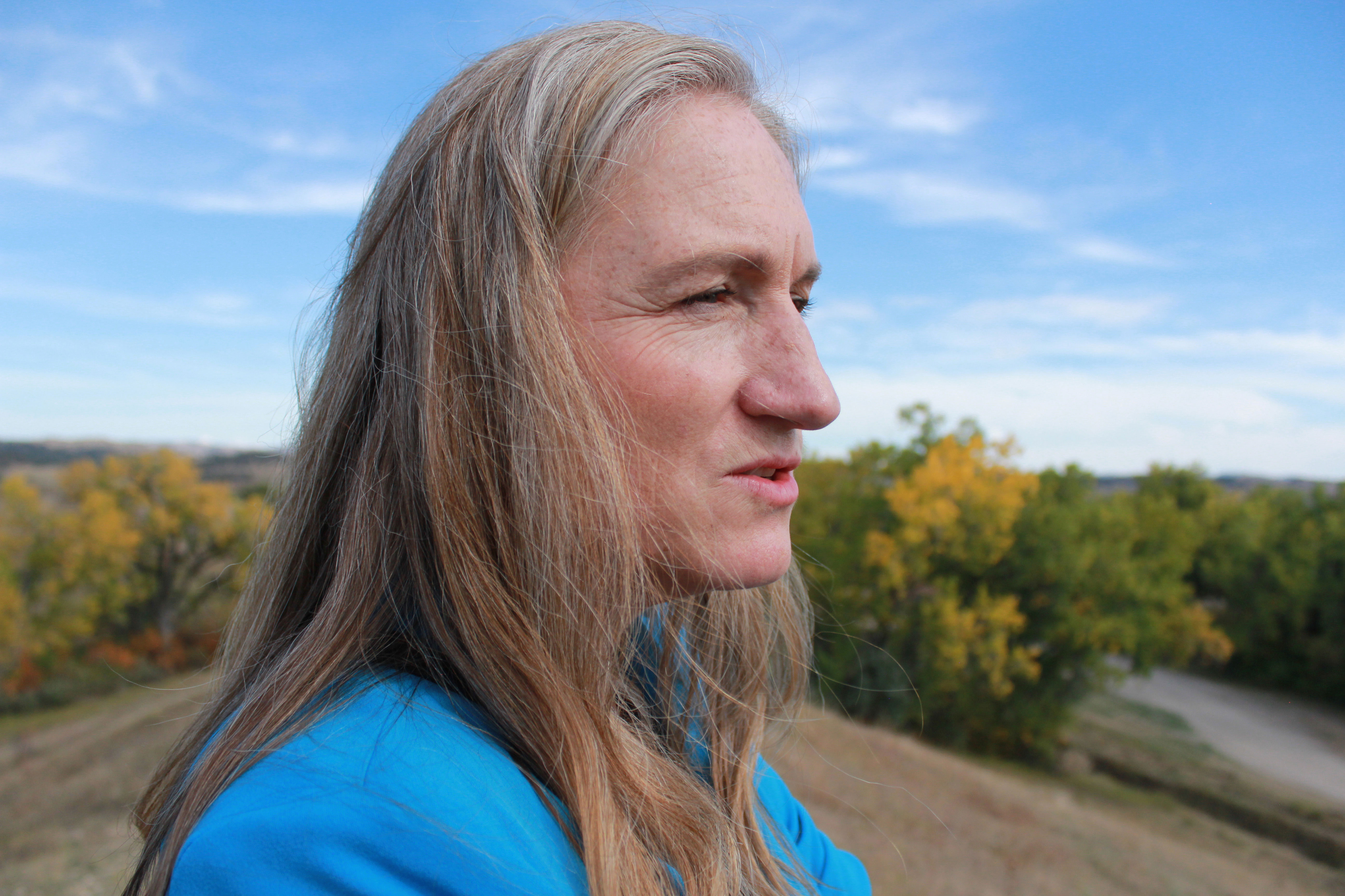 A close-up photo of a woman standing on a hill and looking at something off-camera below the hill. She has long gray and blonde hair, wears a blue top, and is in the middle of speaking.