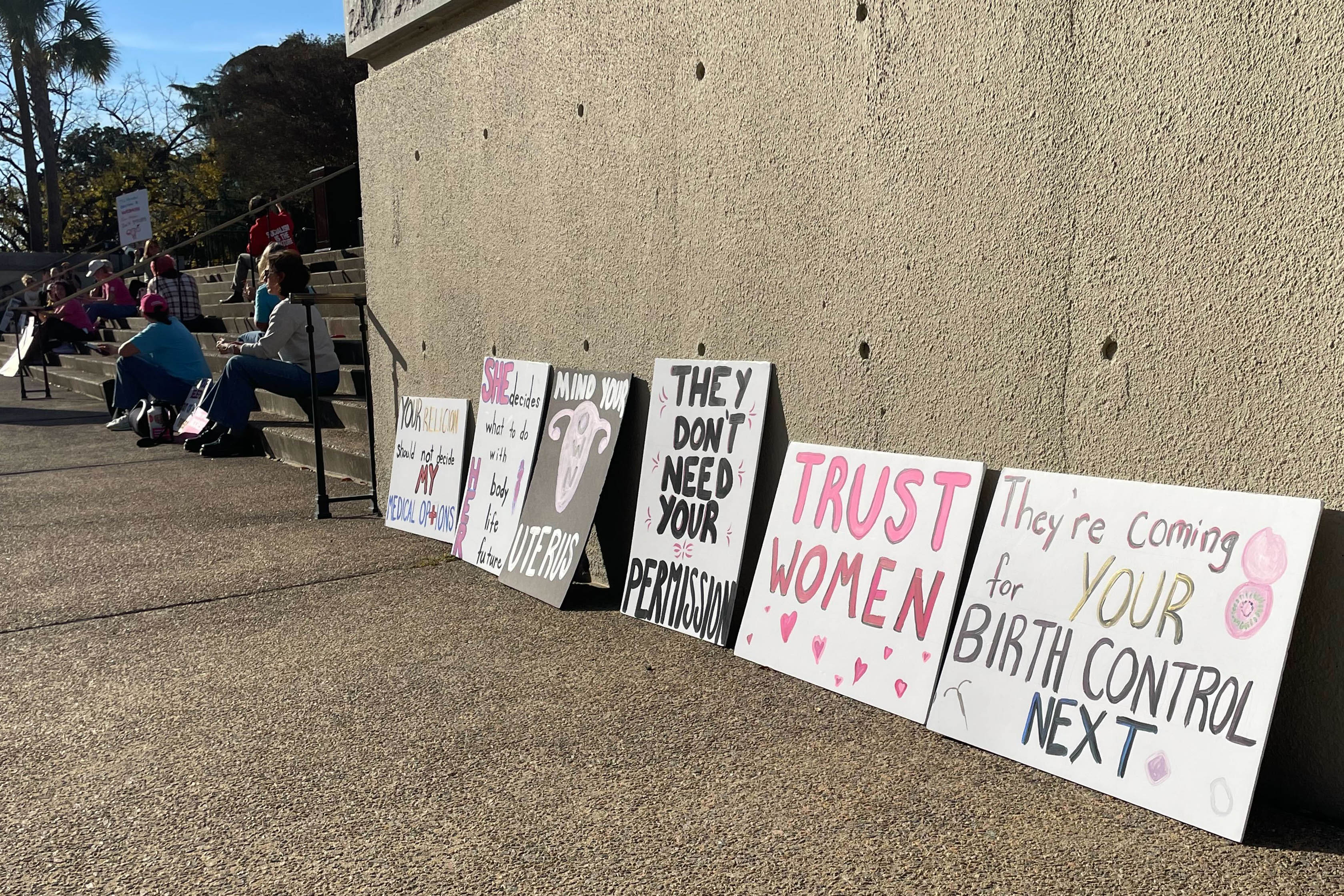 A line of protest signs lean up against a cement wall. They read, "trust women," "they're coming for your birth control next," "they don't need permission," "mind your own uterus," "she decides what to do with her body, life, future."
