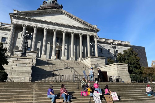 A small group of people sit and stand on the steps of the South Carolina State House.