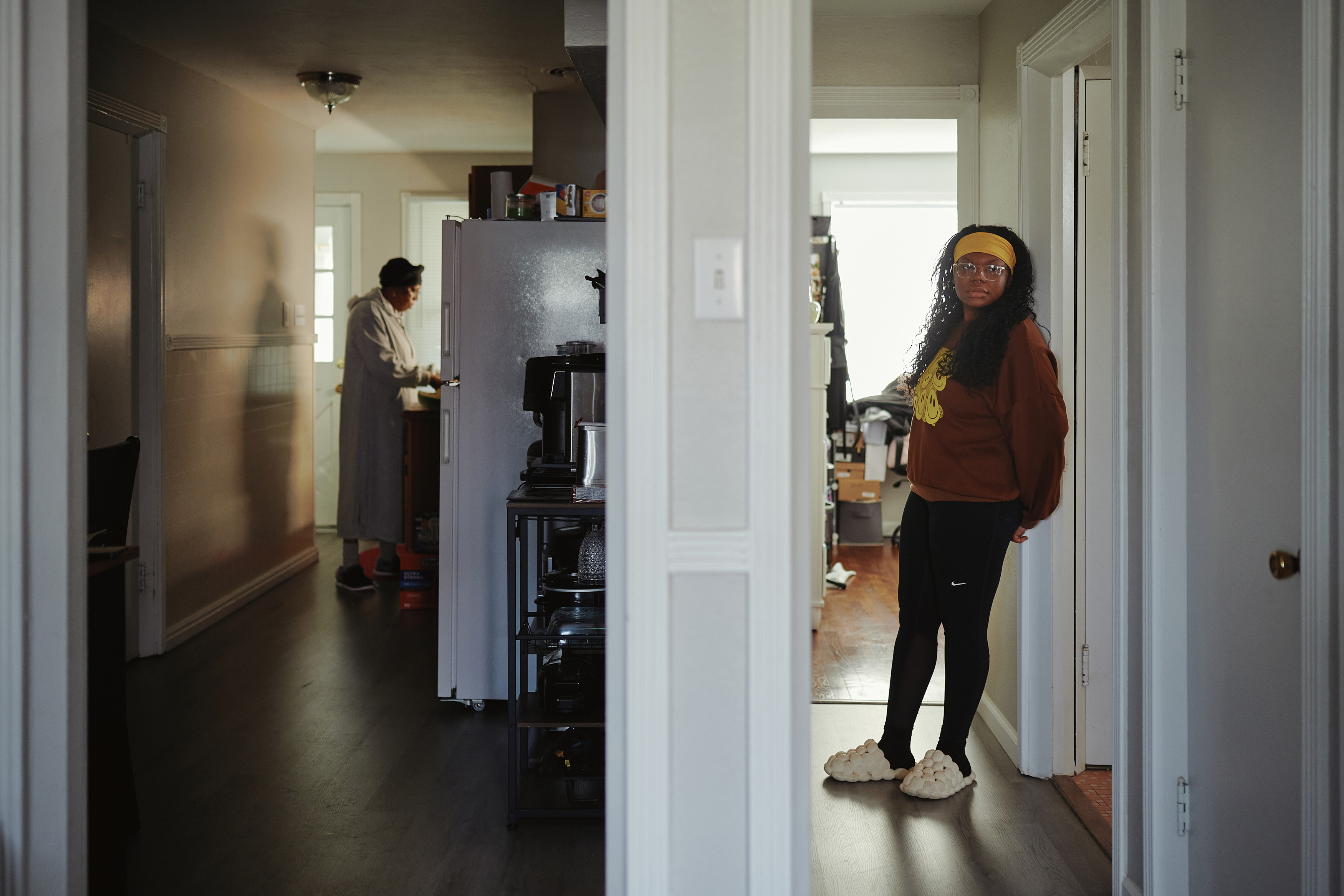 A woman (left) cooks in a kitchen while a woman (right) stands in a hallway looking past the camera