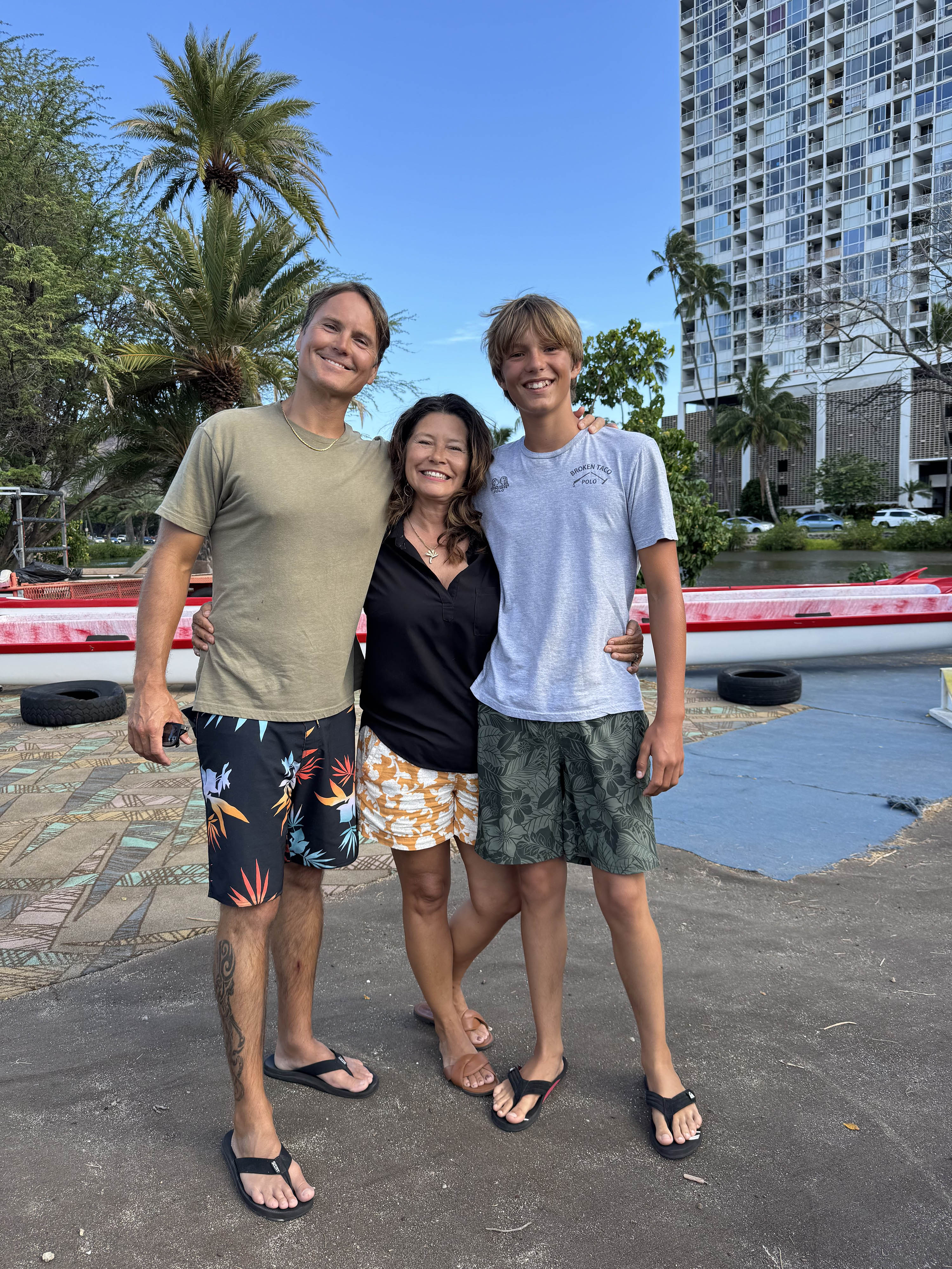 A man, a woman, and a boy pose for a photo with their arms around one another in front of palm trees and a high-rise building