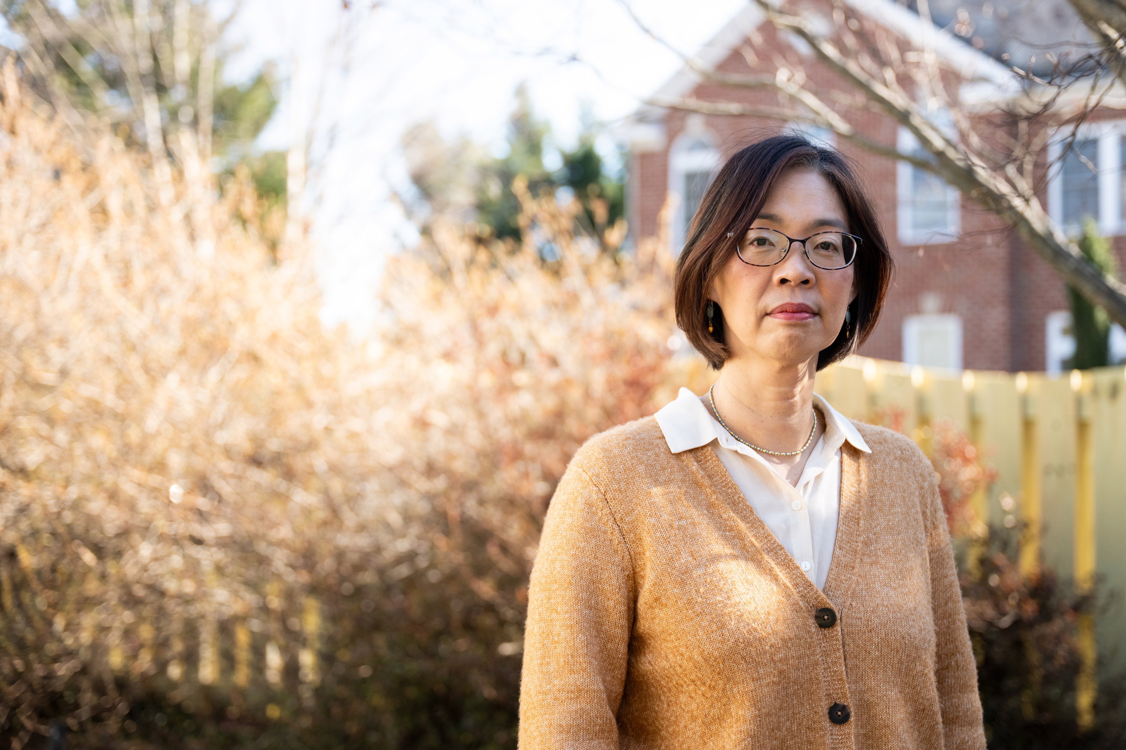 Sylvia Choi stands by a fence in her backyard. Shrubbery and a building are seen behind her.