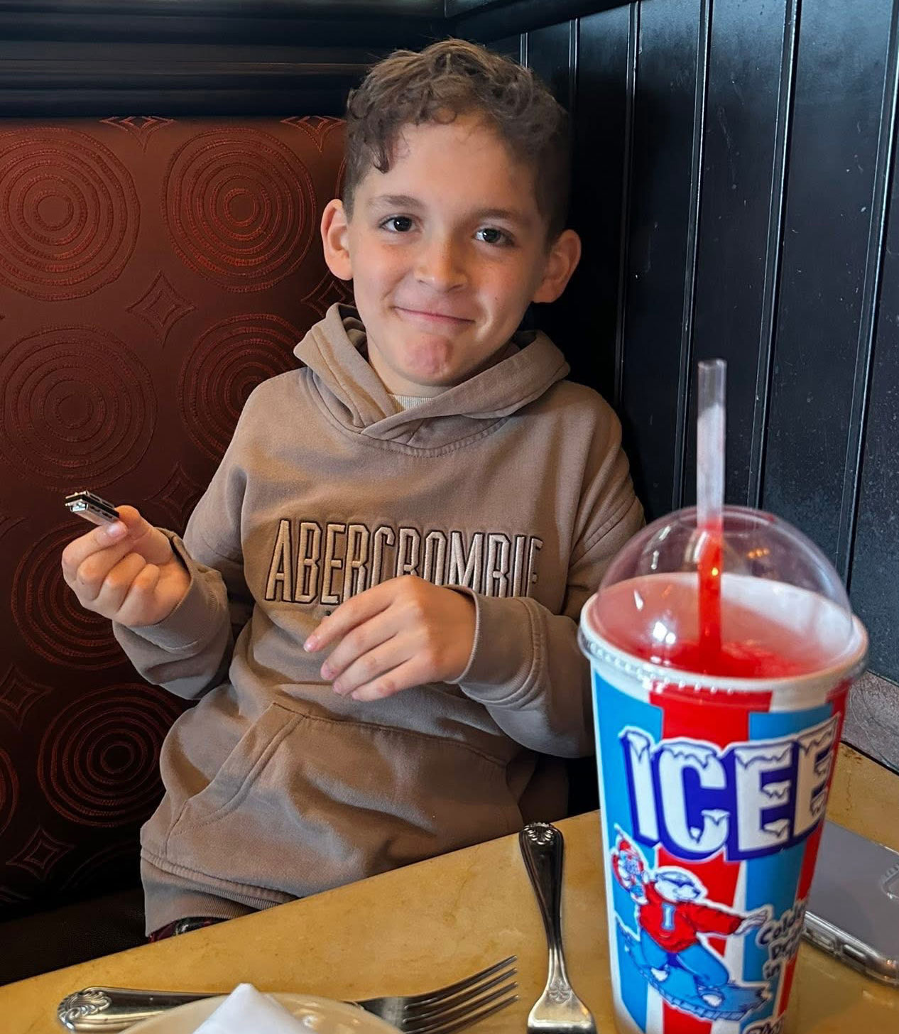 Jonah Reynolds smiles while sitting in a restaurant booth. He holds a miniature harmonica. A cup of red Icee slushie is on the table in front of him.