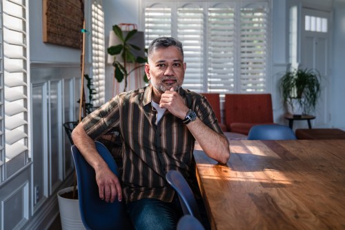 A South Asian man sits by the window at a table. He rests his elbow on the table with his hand on his chin. He looks contemplative.