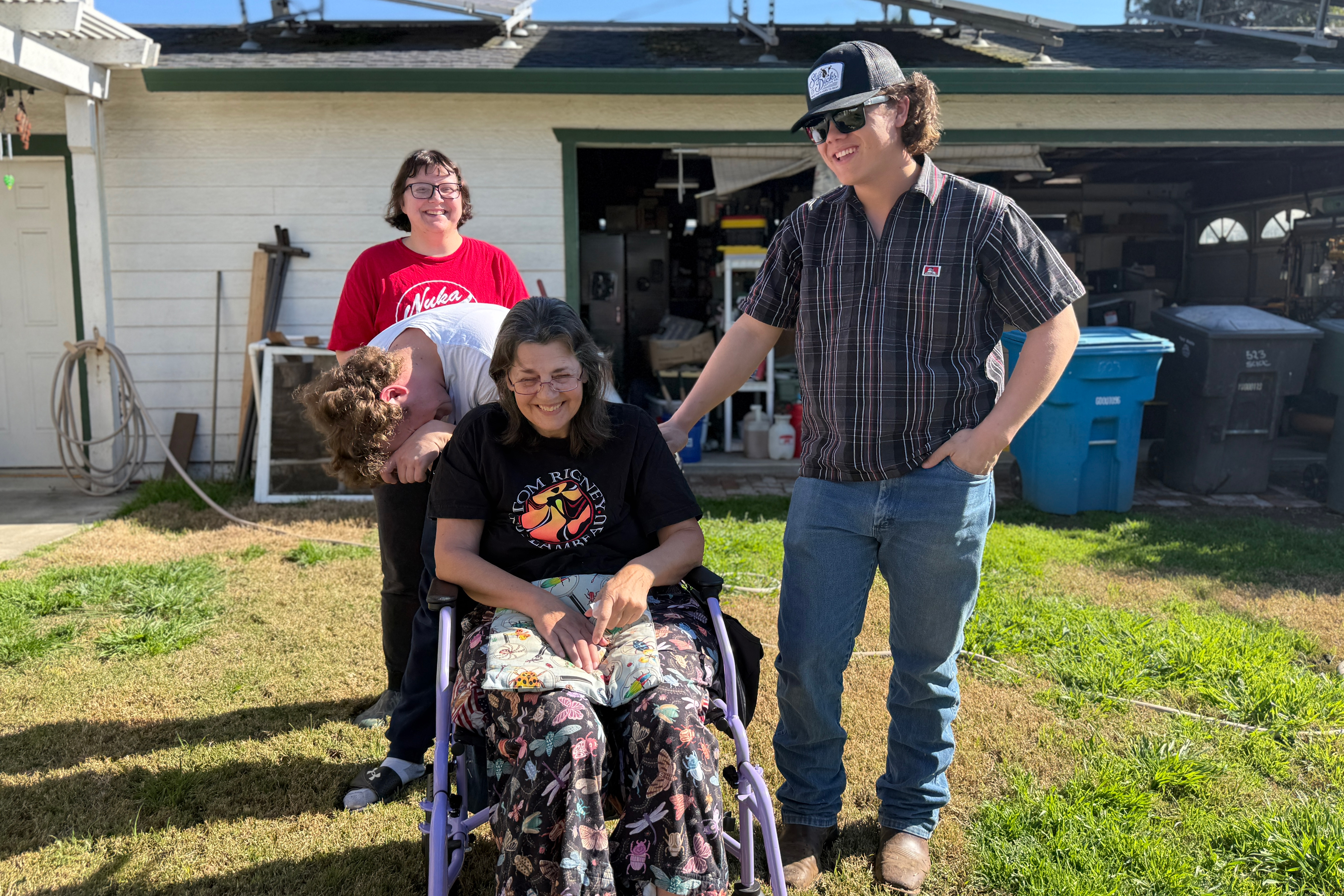A photo of Jean Franklin posing outside her house in her wheelchair. She is surrounded by three younger people: two of her sons and one of her son's girlfriend.