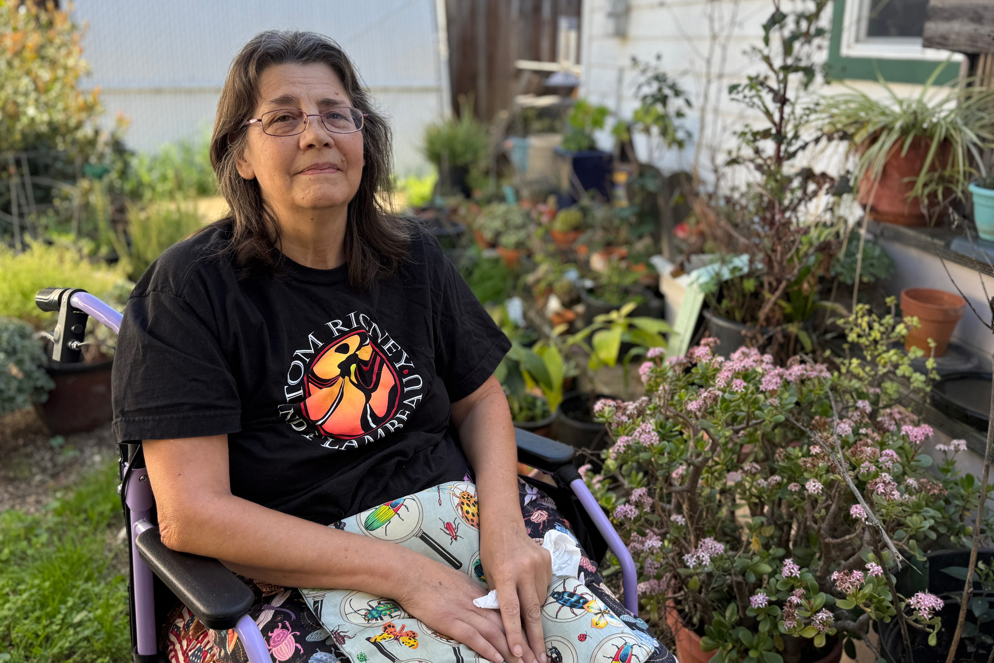 Jean Franklin poses for a photo in her wheelchair. She is seen sitting next to many potted plants outside.