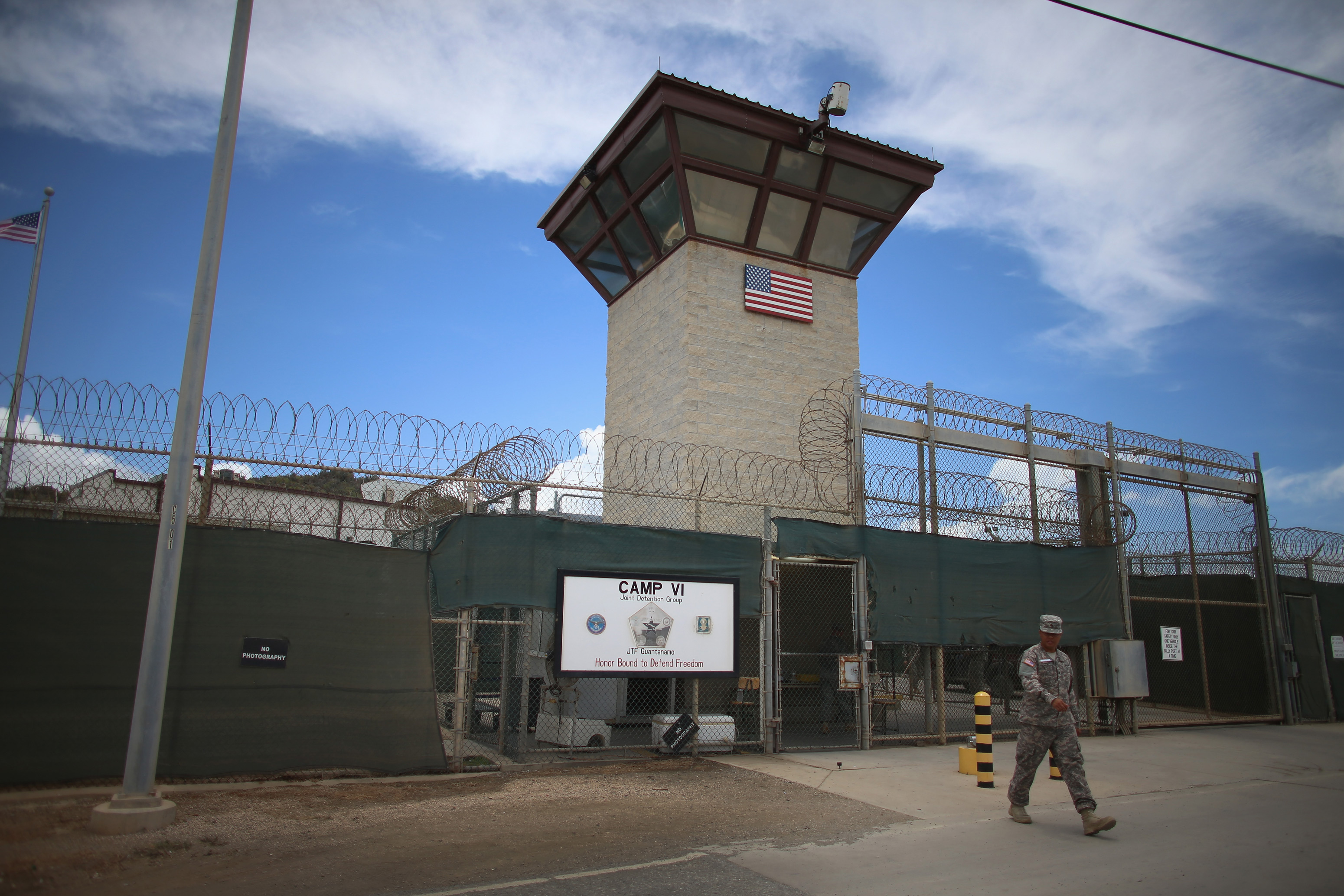 An exterior view of Camp 6. A guard tower is seen with barbed-wire fences. A man in military uniform is seen walking in the foreground.