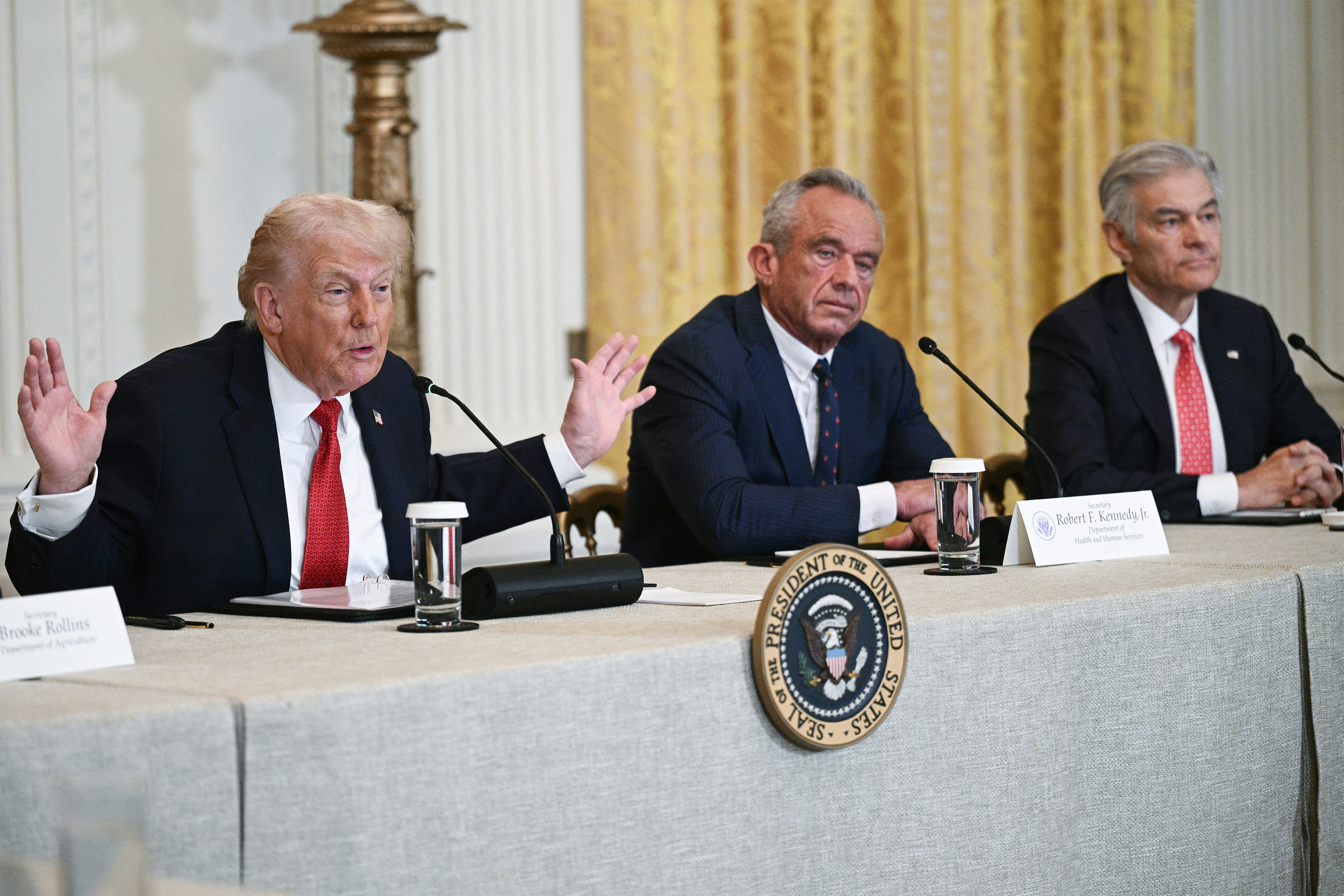 A photo of a White House event: President Trump sits at a table. To the right of him is Robert F. Kennedy Jr. Mehmet Oz sits to Kennedy's right. All three men have microphones across from them.