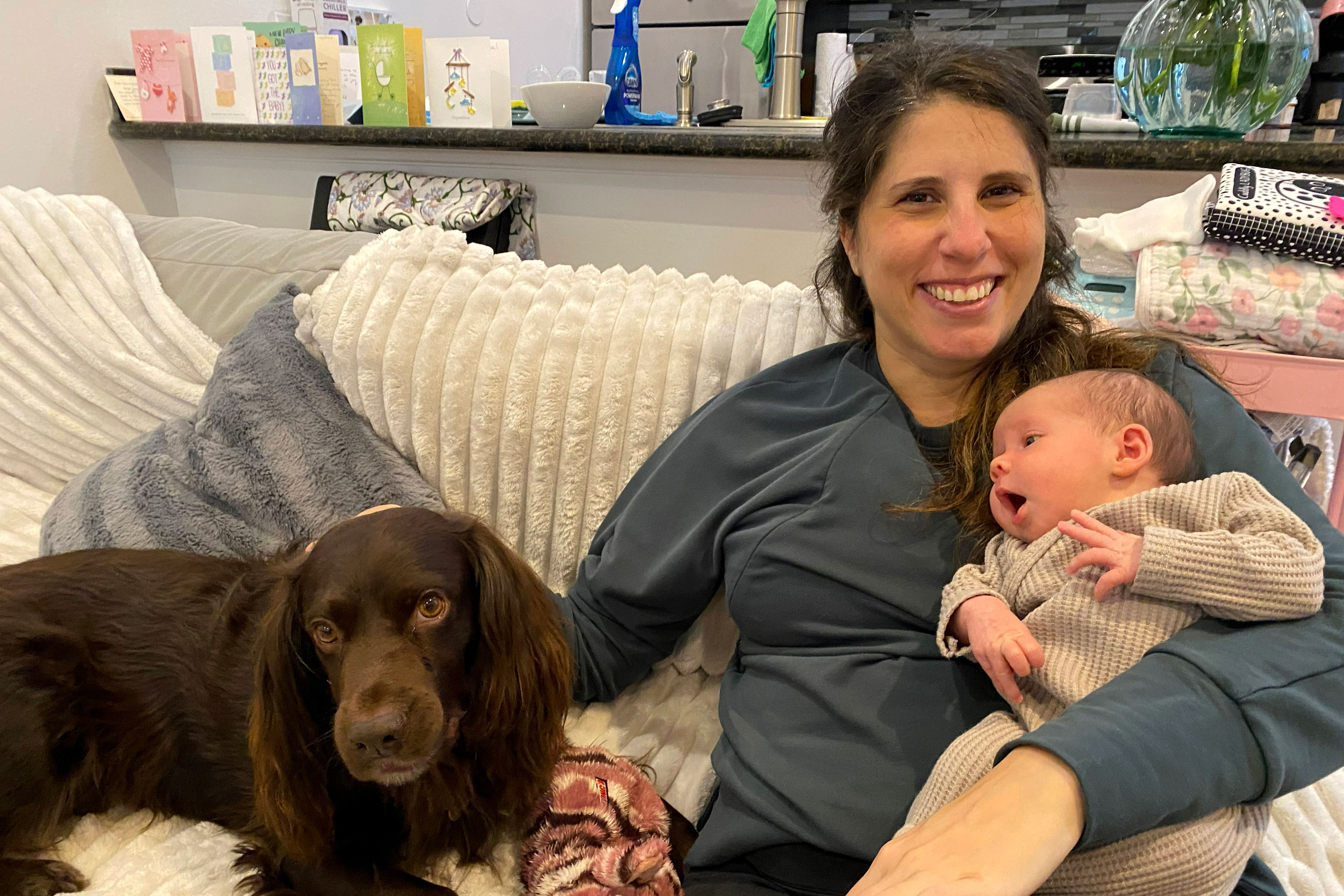 A woman sits on a couch with a young baby in her left arm and her right hand petting a brown dog sitting beside her. She smiles at the camera.