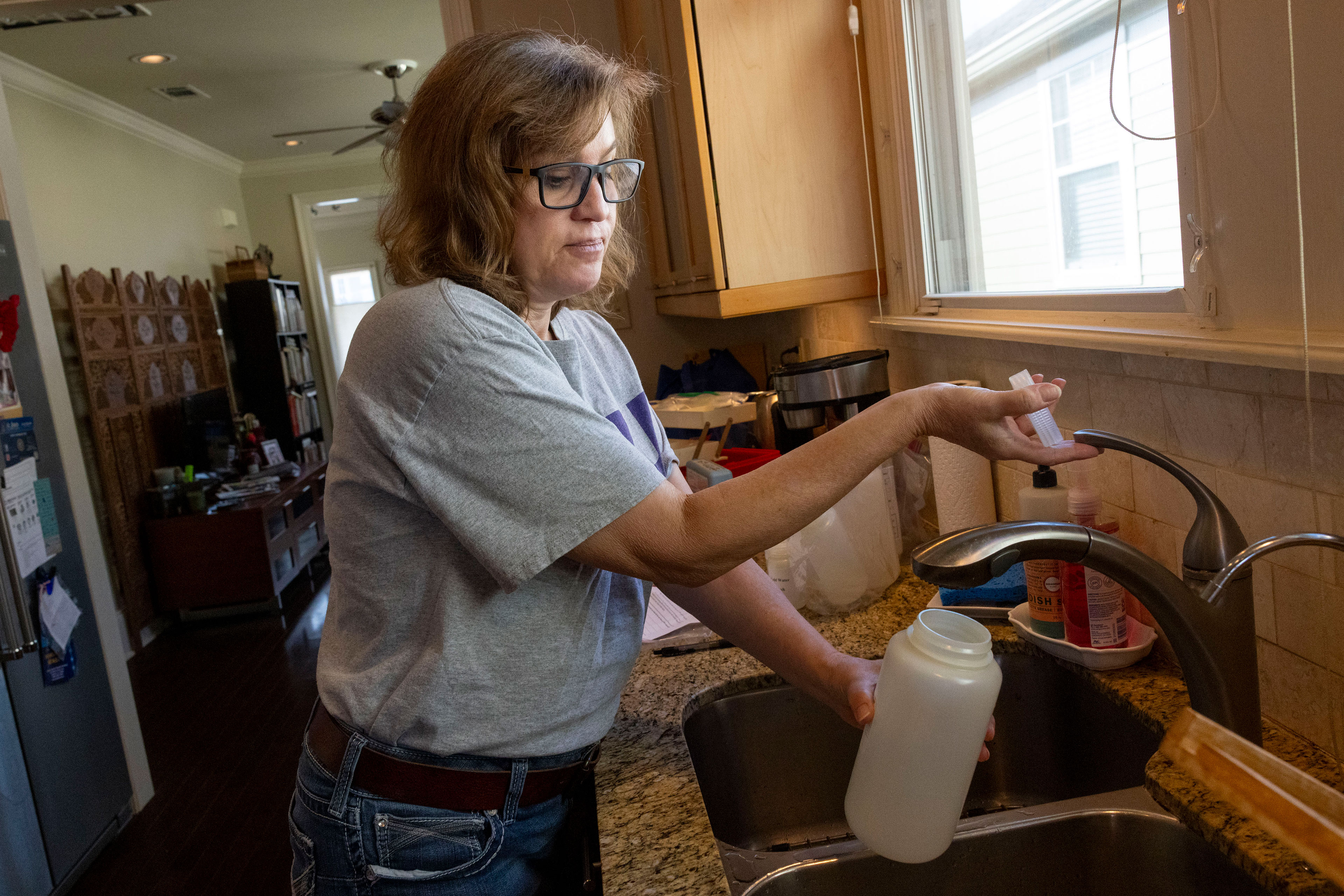 A woman in a gray t-shirt fills a container with water from a kitchen faucet.