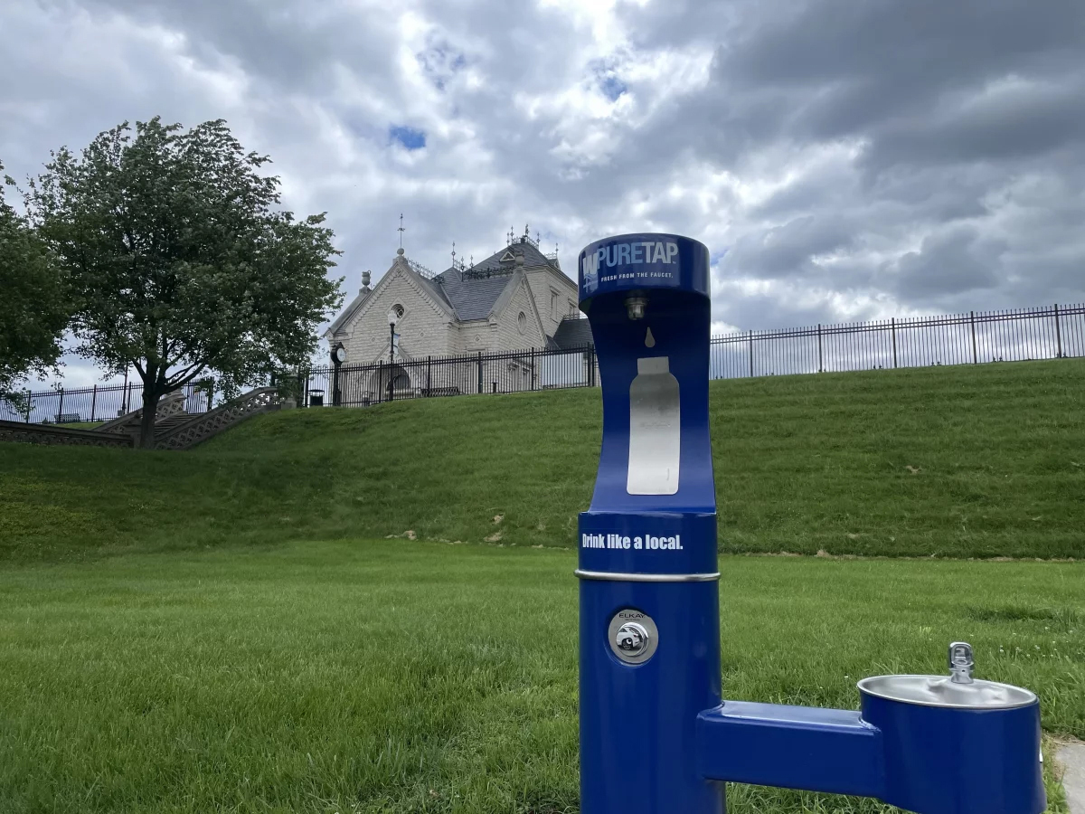 A blue water fountain and water bottle fill station stands near a grassy field
