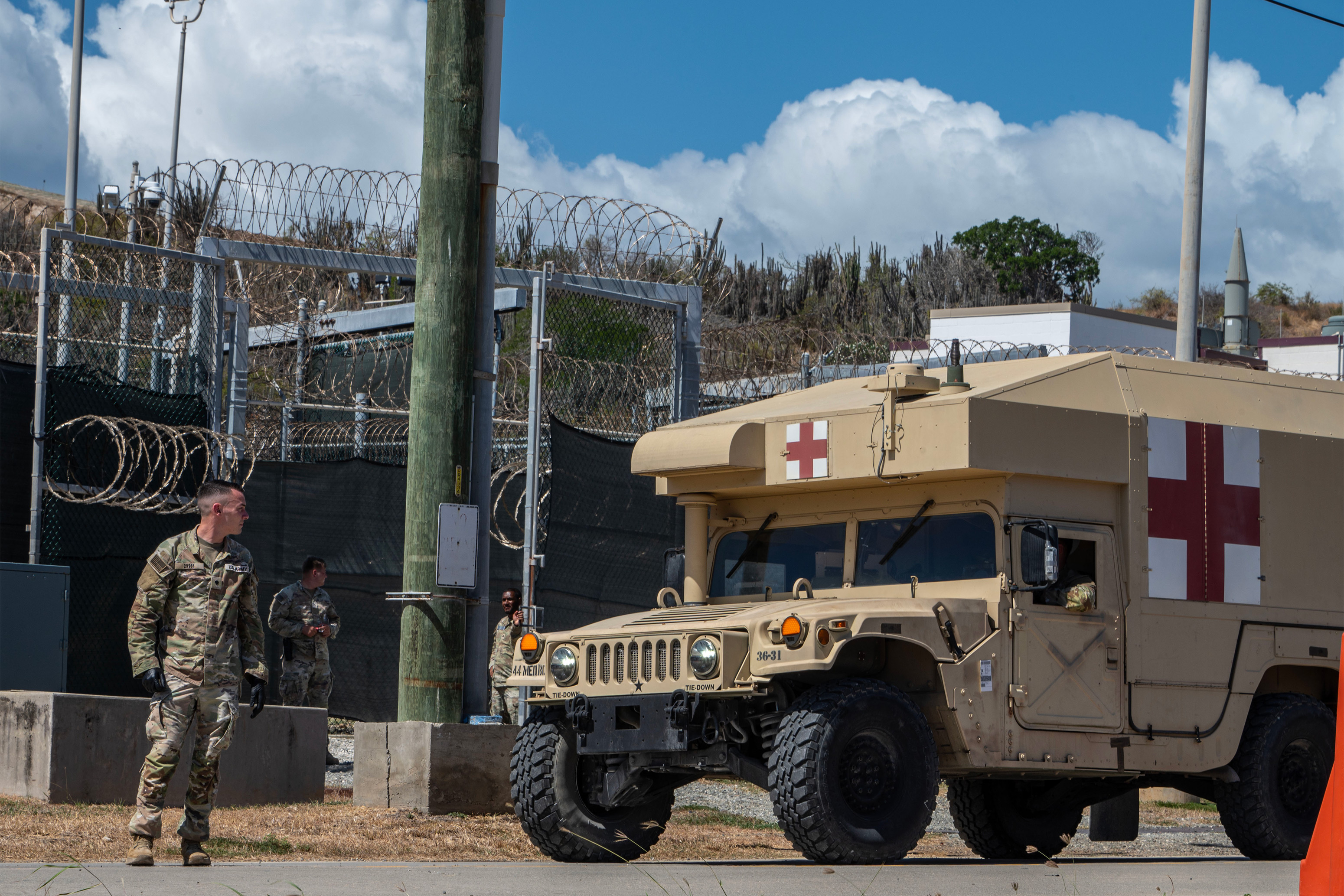 A military vehicle with a red cross symbol painted on it approaches three military members. A barbed-wire fence is behind them.
