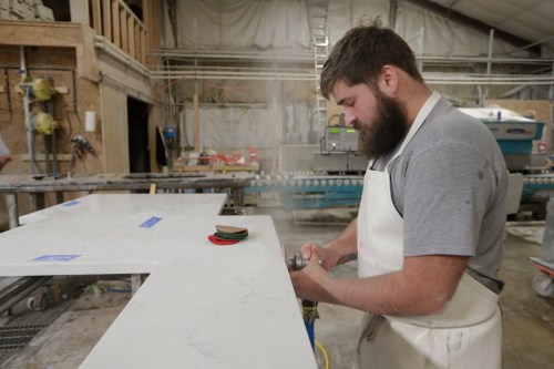 A man works with a polishing tool on the edge of a countertop. Dust can be seen flying in the air around the tool. Some of the dust has settled in the man's hair.