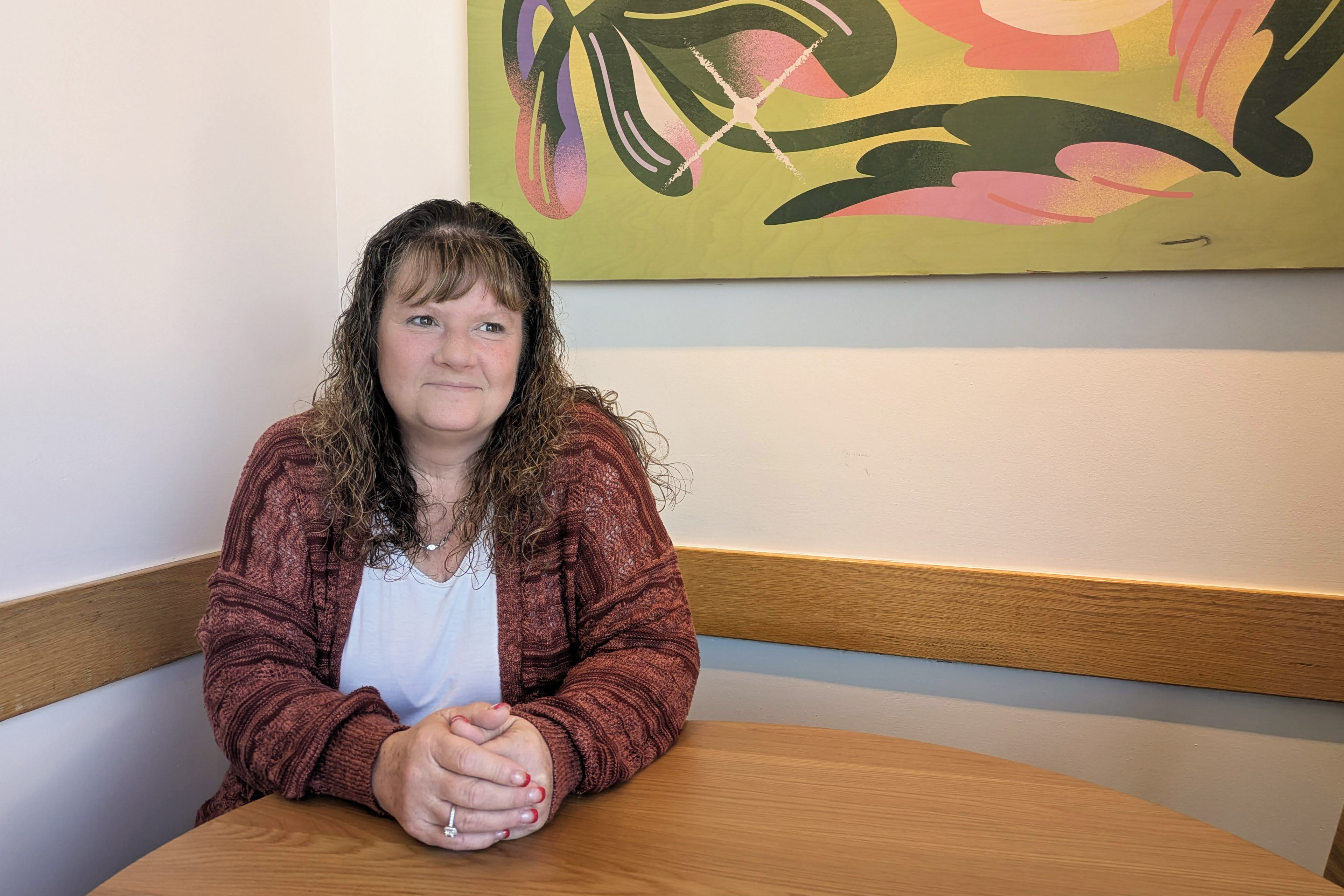 A woman sits at a table indoors. A pink and green abstract painting in seen on the wall behind her.