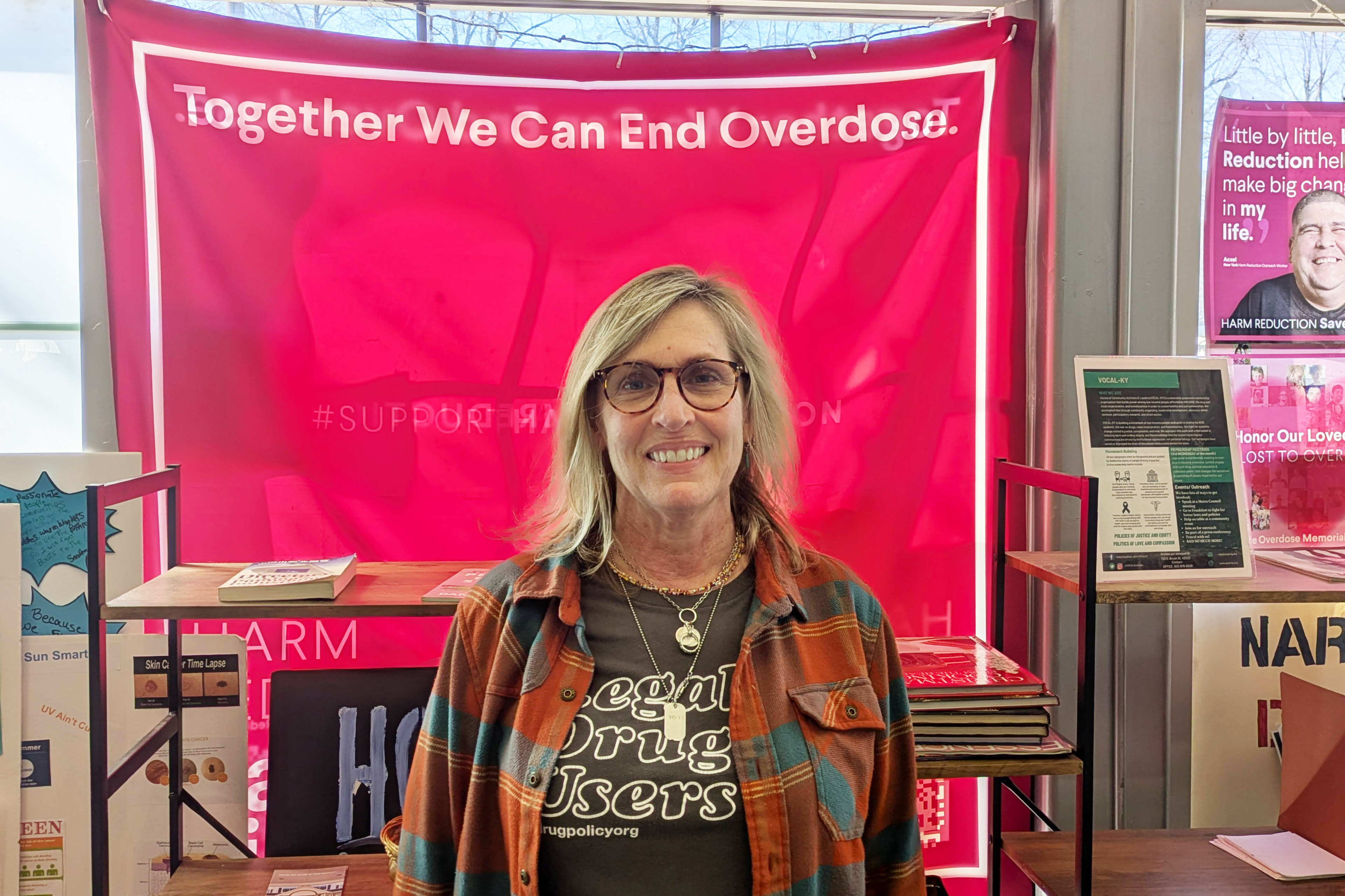 A woman stands indoors. Behind her is a pink banner that reads, "Together We Can End Overdose."