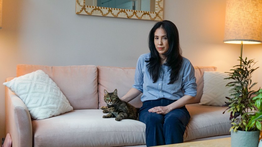 A woman sits on a couch with a tabby cat beside her. Her hand gently rests beside her cat.