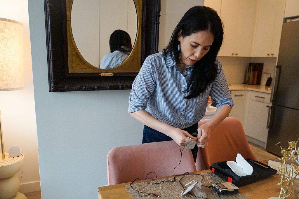 A woman leans over a dining room table, where she handles a medical device.