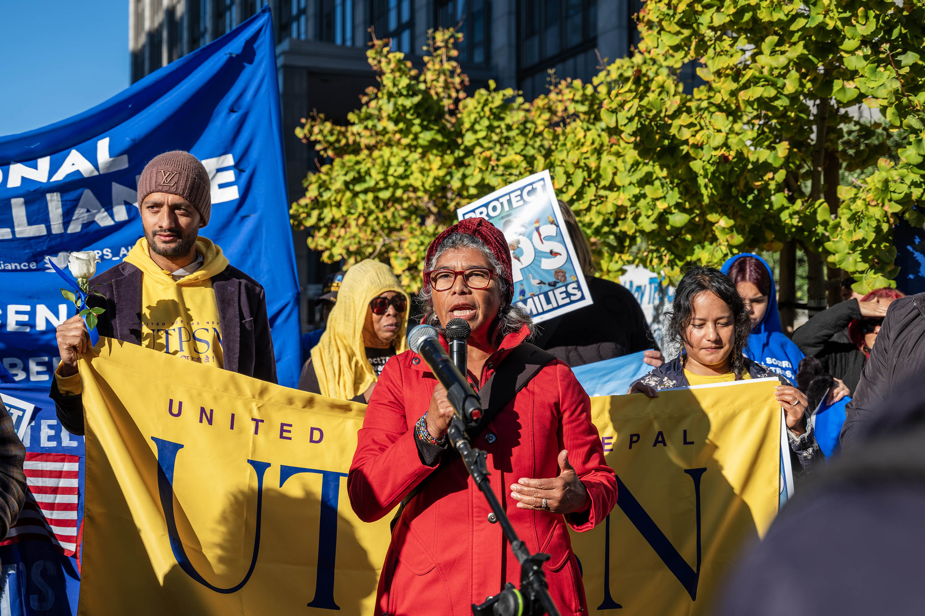 A woman in a red jacket holds a microphone as she speaks to a crowd of people. Behind her, protesters hold a banner and signs.