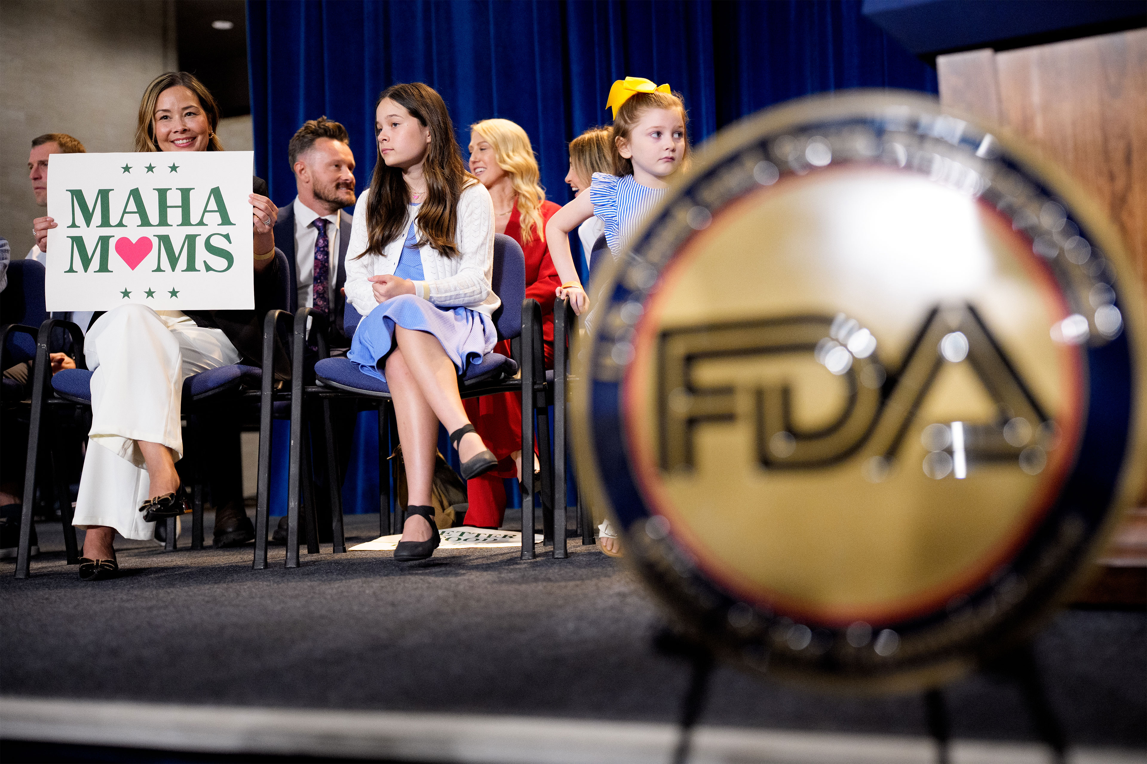 A woman holds a sign that says "MAHA Moms" as she sits on stage at a news conference at the Health and Human Services Department in Washington. Two young girls sit beside her. The insignia for the FDA is seen blurred in the foreground.