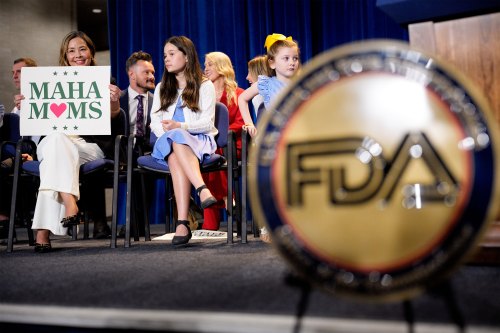 A woman holds a sign that says "MAHA Moms" as she sits on stage at a news conference at the Health and Human Services Department in Washington. Two young girls sit beside her. The insignia for the FDA is seen blurred in the foreground.