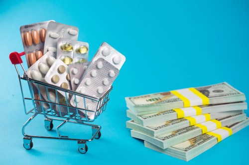 A photo of a mini shopping cart filled with pills beside a stack of money against a blue background.