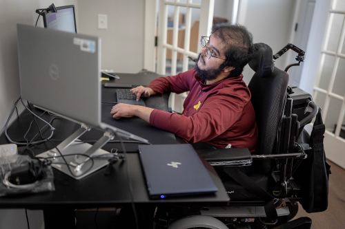 A man with muscular dystrophy works at a desk with multiple computer monitors.