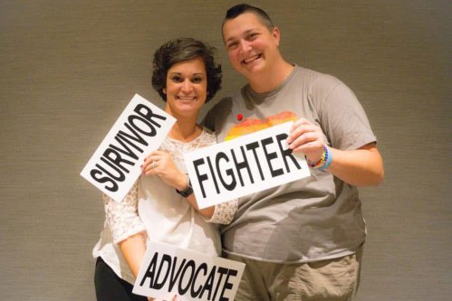 Two people pose for a photo together, smiling. The person on the left holds signs that read, "Survivor" and "Avocate." The person the right holds a sign that reads, "Fighter."