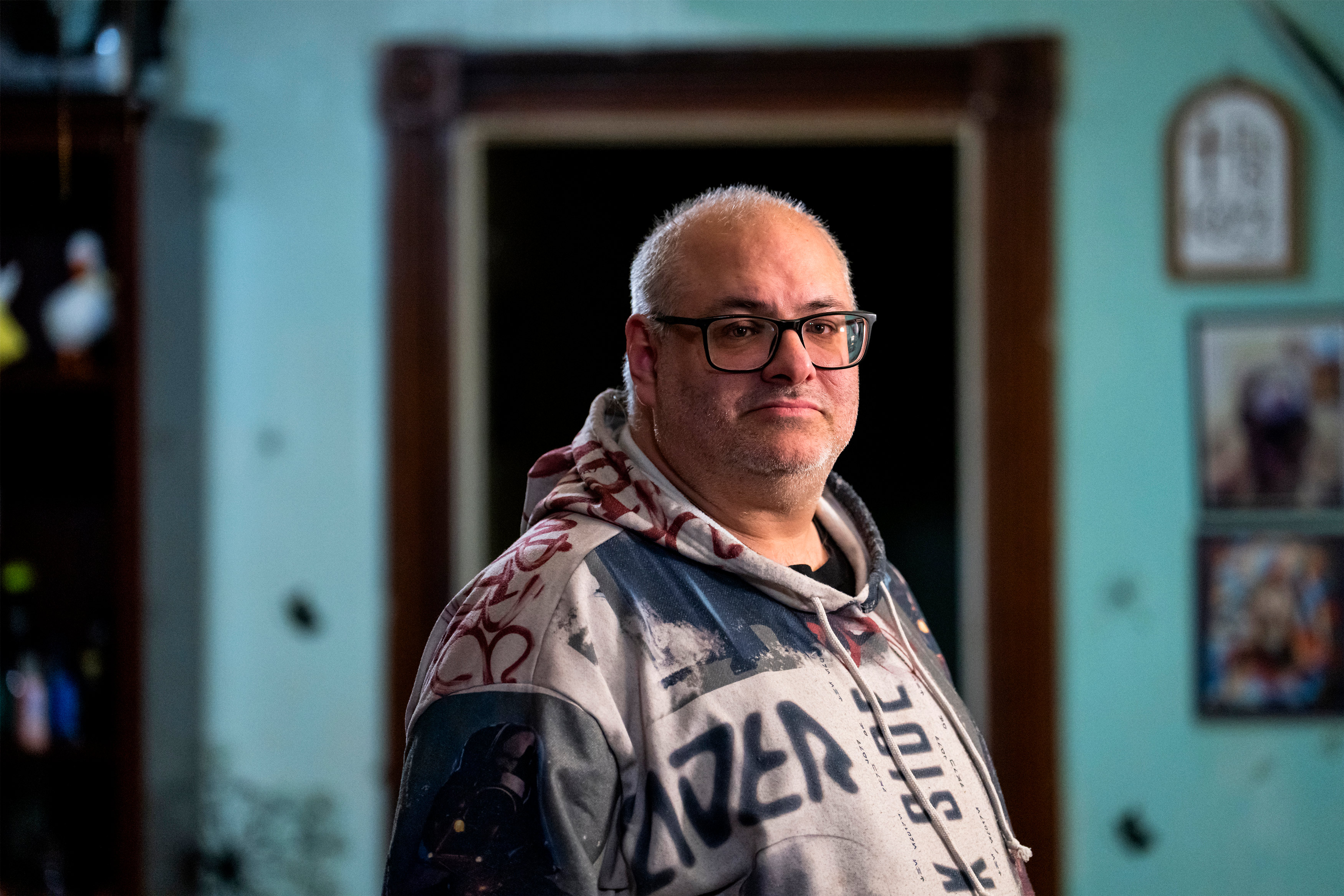 A man stands indoors for a portrait. He is wearing a hoodie,