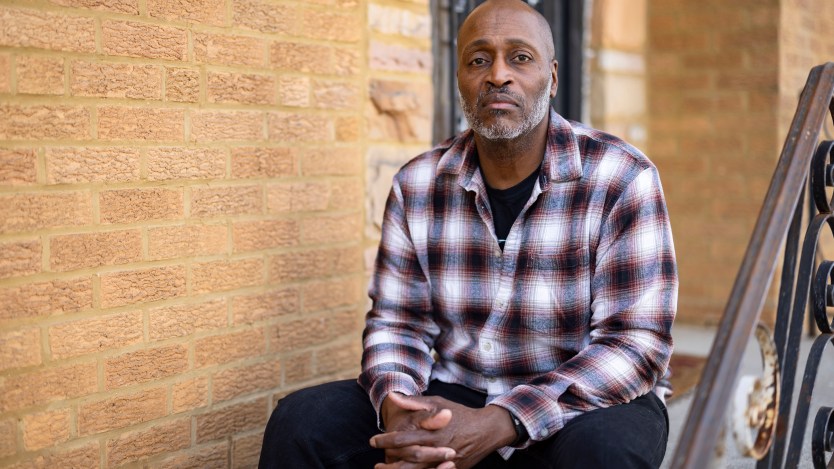 A man wearing a plaid button-up shirt sits on the front step of a home for a portrait