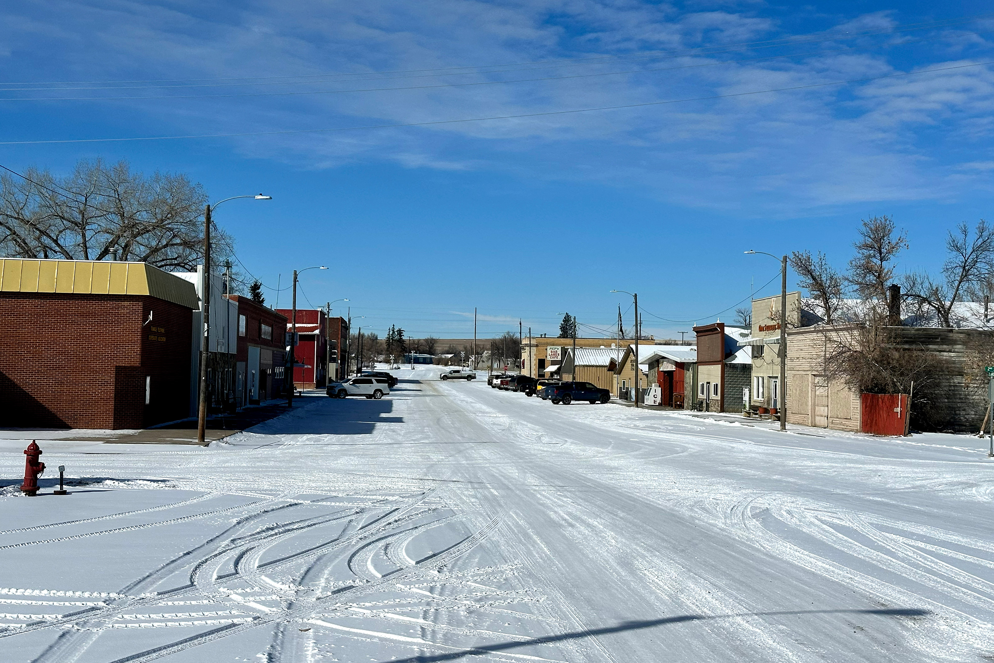 A snow-covered street in a rural town with shops lining it. A few cars are parked in front of the businesses.