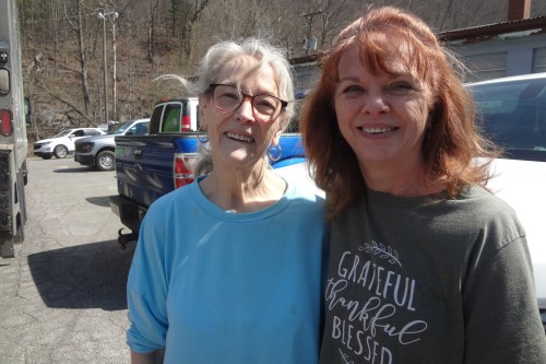 Two older women stand smiling side by side in a parking lot on a sunny day.