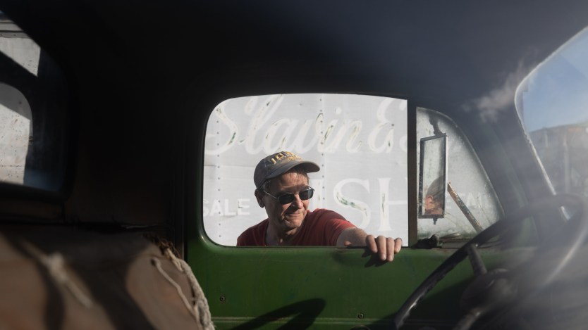 A man wearing a red shirt, a baseball cap and sunglasses rests his hand on the open window of a truck