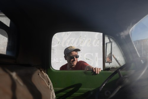 A man wearing a red shirt, a baseball cap and sunglasses rests his hand on the open window of a truck