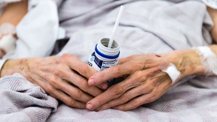 A close-up photograph of a frail senior's hands holding a bottled protein shake with a straw in it. The person wears a hospital gown, is covered in a blanket, and has bandages on one hand and their arms.