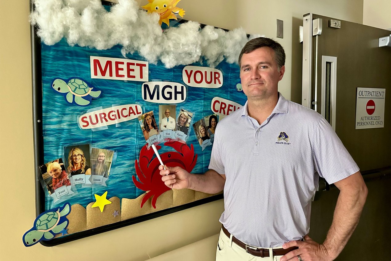 A man stands indoors. He stands next to a decorated bulletin board. It reads, "Meet your MGH surgical crew." Below it are sets of photos of hospital staff. The man points with a pen to a photo of a woman.