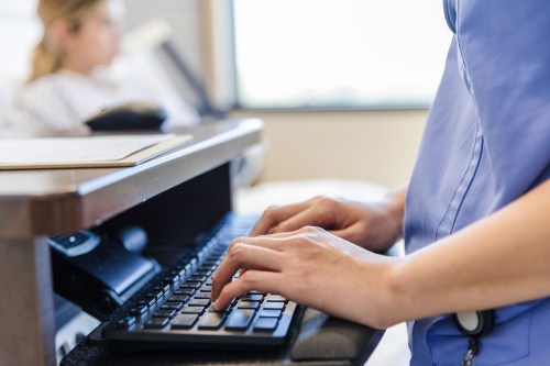 An unidentifiable nurse types at a computer. A patient is visible, but blurry, in the background.