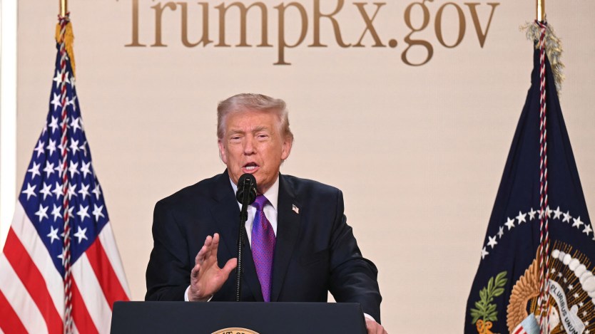 President Trump speaks at a podium. The American flag is to the left of him. A flag with the presidential seal is to the right of him. Behind him is a sign that reads, "TrumpRx.gov."
