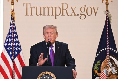 President Trump speaks at a podium. The American flag is to the left of him. A flag with the presidential seal is to the right of him. Behind him is a sign that reads, "TrumpRx.gov."