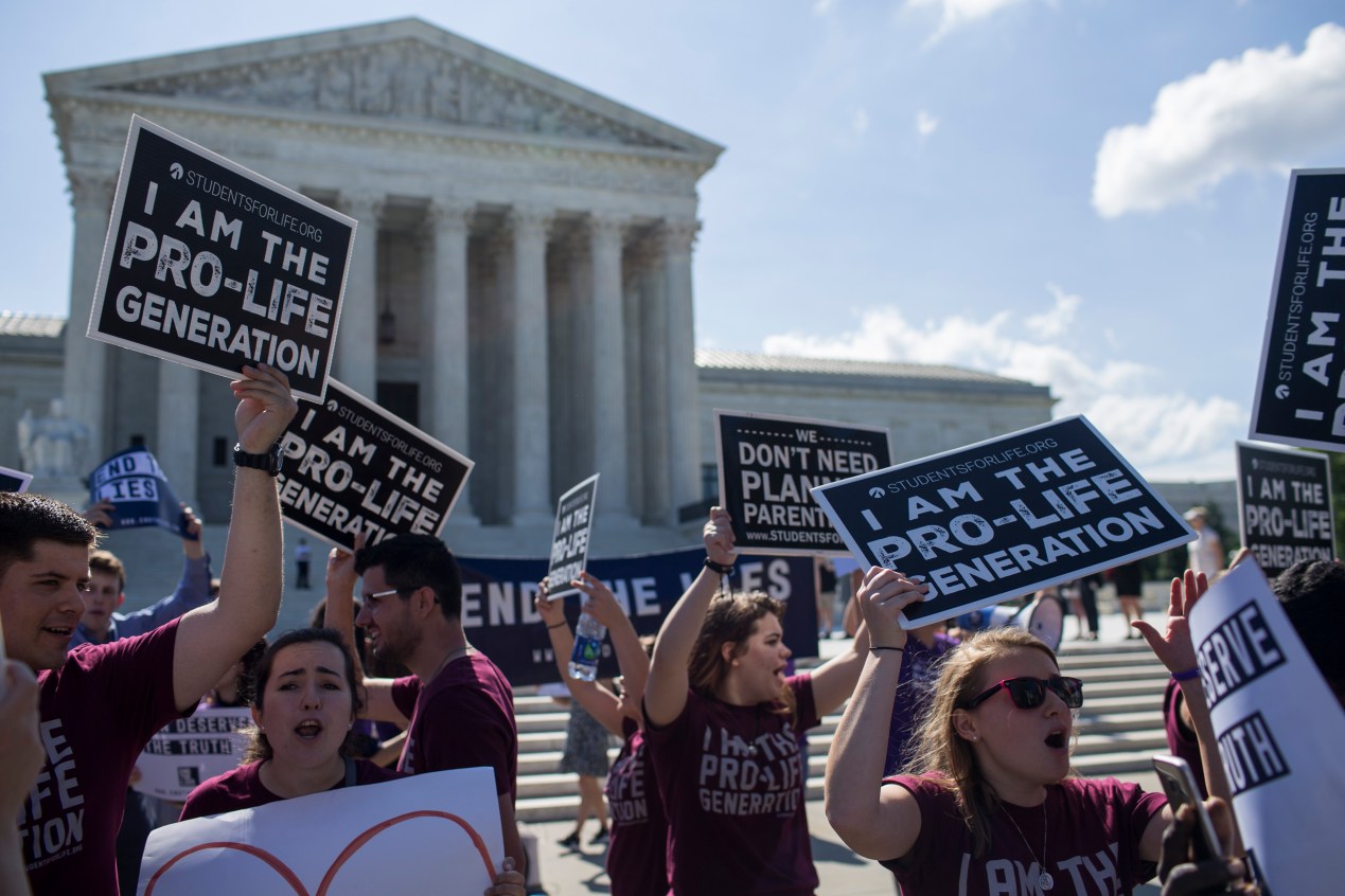 A photo of anti-abortion protesters in front of the Supreme Court. They hold signs reading, "I am the pro-life generation," and "We don't need Planned Parenthood."