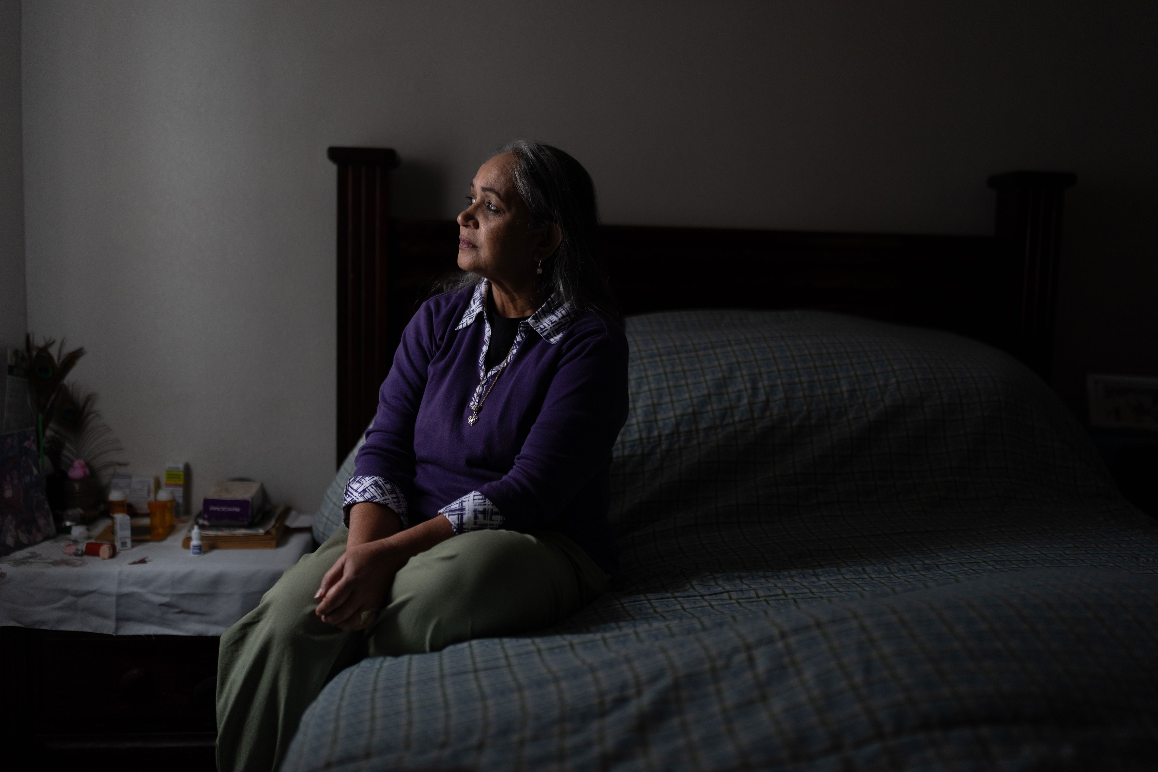 A woman in a bedroom lit only by some light from a window sits on a bed and looks out that window contemplatively.