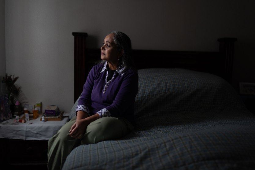 A woman in a bedroom lit only by some light from a window sits on a bed and looks out that window contemplatively.