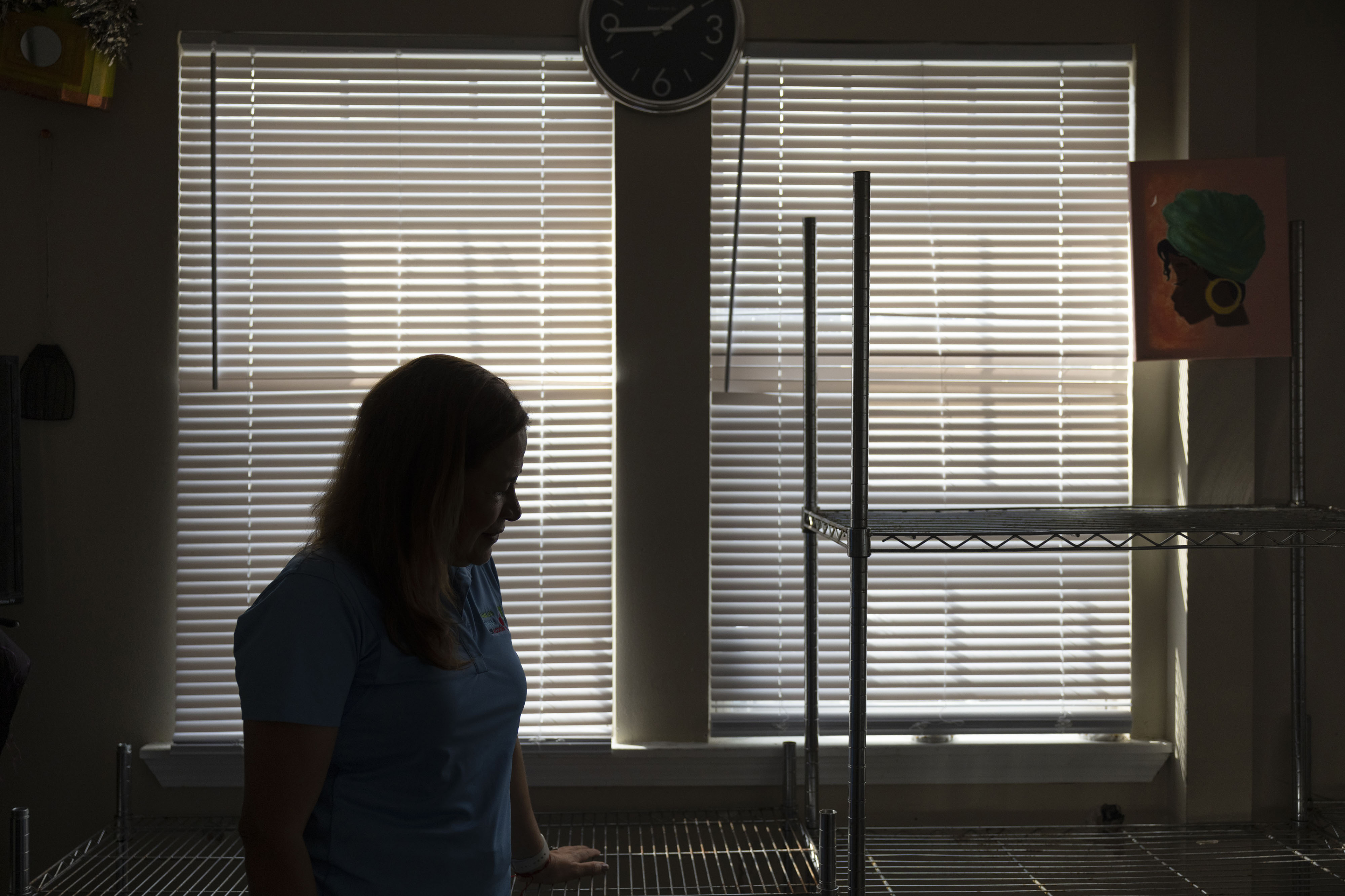 A woman stands near some shelves indoors and is in silhouette against two windows with blinds drawn.
