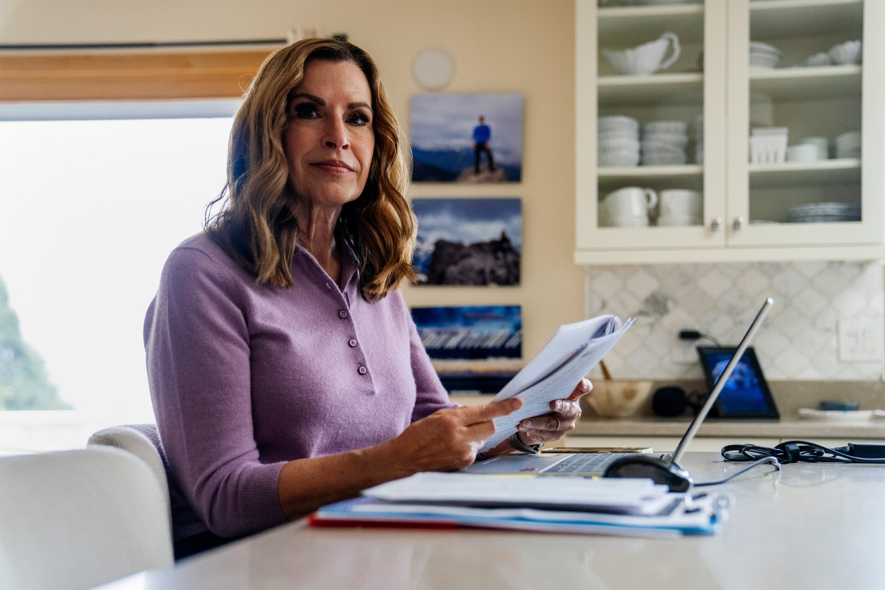 Jan Anderson sits at a kitchen island counter. A laptop and paperwork is in front of her. She holds paperwork in her hands.