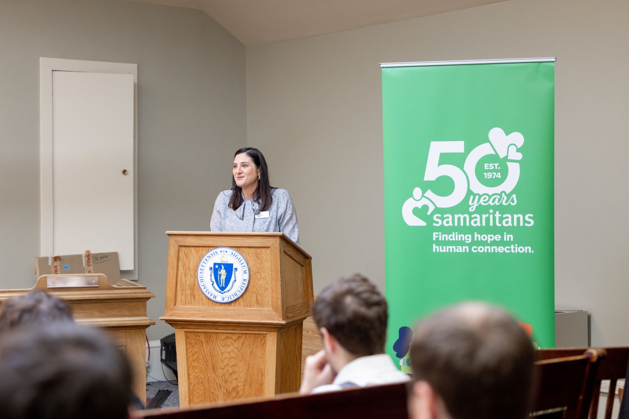 A woman with straight brown hair and wearing a light blue blazer stands at a podium as she speaks to a small audience.