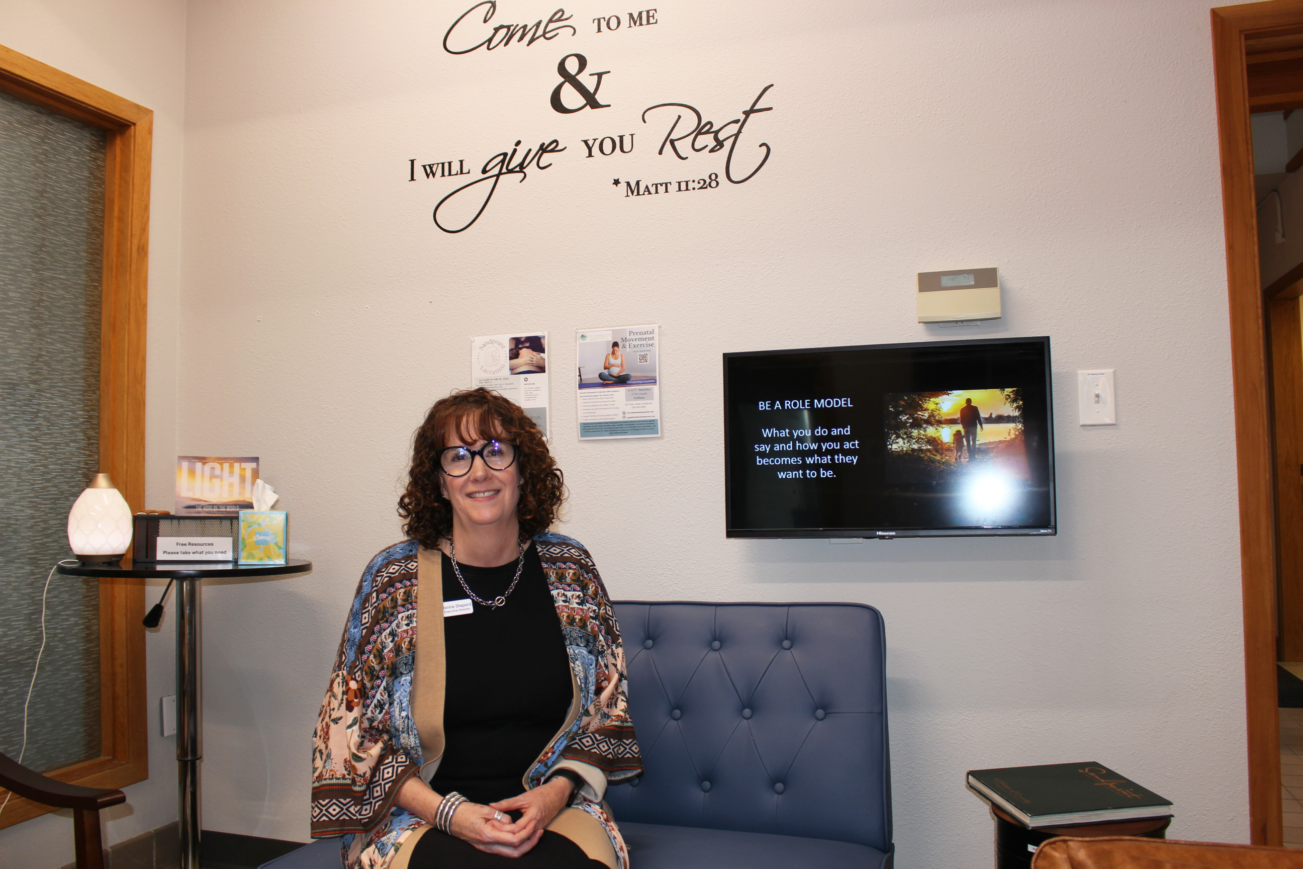 A woman sits in a chair at a crisis pregnant center. The wall behind her has Biblical scripture written on it: "Come to me & I will give you rest." A TV monitor next to her shows a slide that reads, "Be a role model. What you do and say and how you act becomes what they want to be."